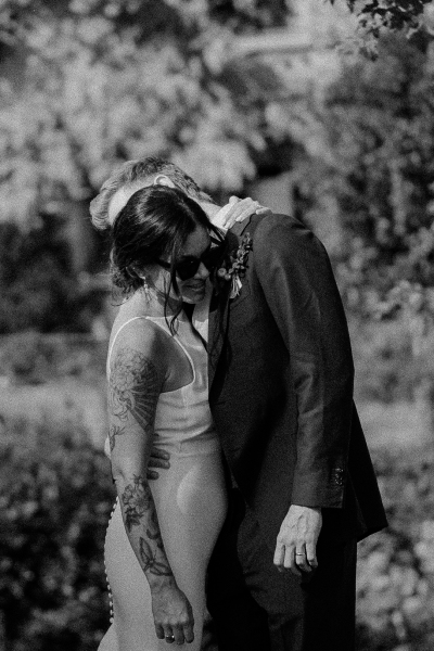 Bride in a sleeveless dress and groom sharing a personal moment outdoors, both smiling, with the bride wearing sunglasses and showcasing a tattoo on her arm.